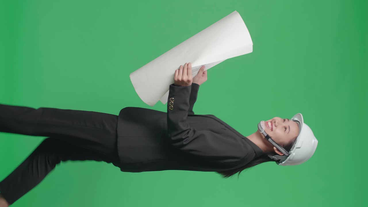 Side View Of Smiling Asian Female Engineer Looking At Blueprint Then Looking Around And Shaking Her Head While Walking In The Green Screen Studio