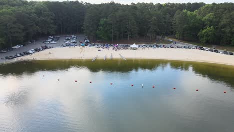 drone camera flying across a lake beach while people stand on the beach preparing for a dragon boat race