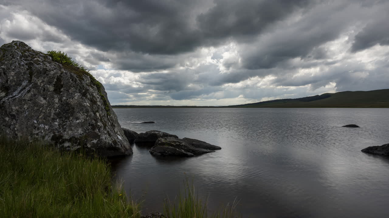 lapso de tiempo del lago con hierba y grandes rocas en primer plano en un oscuro día de verano nublado en el paisaje rural de irlanda