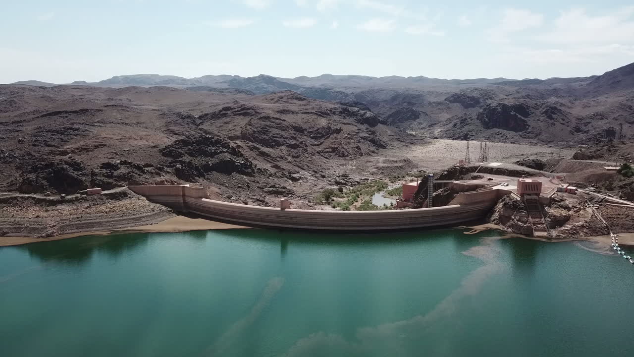 Panoramica aerea 4K completa di un imponente complesso di dighe. La ripresa cattura la parete curva in cemento, il bacino turchese e gli edifici delle infrastrutture tecniche all'interno di un terreno desertico accidentato. Ideale.