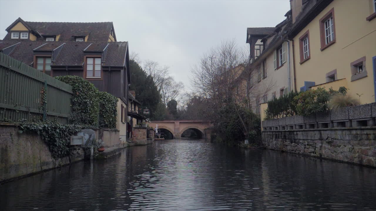 En rolig engel af en sidekanal i Colmar, Frankrig, med roligt vand, en enkel stenbro og historiske huse, hvoraf det ene har efeu og et træhegn