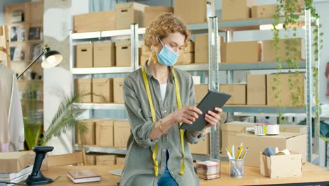 young caucasian designer woman in facial mask and typing on tablet at office in clothing shop