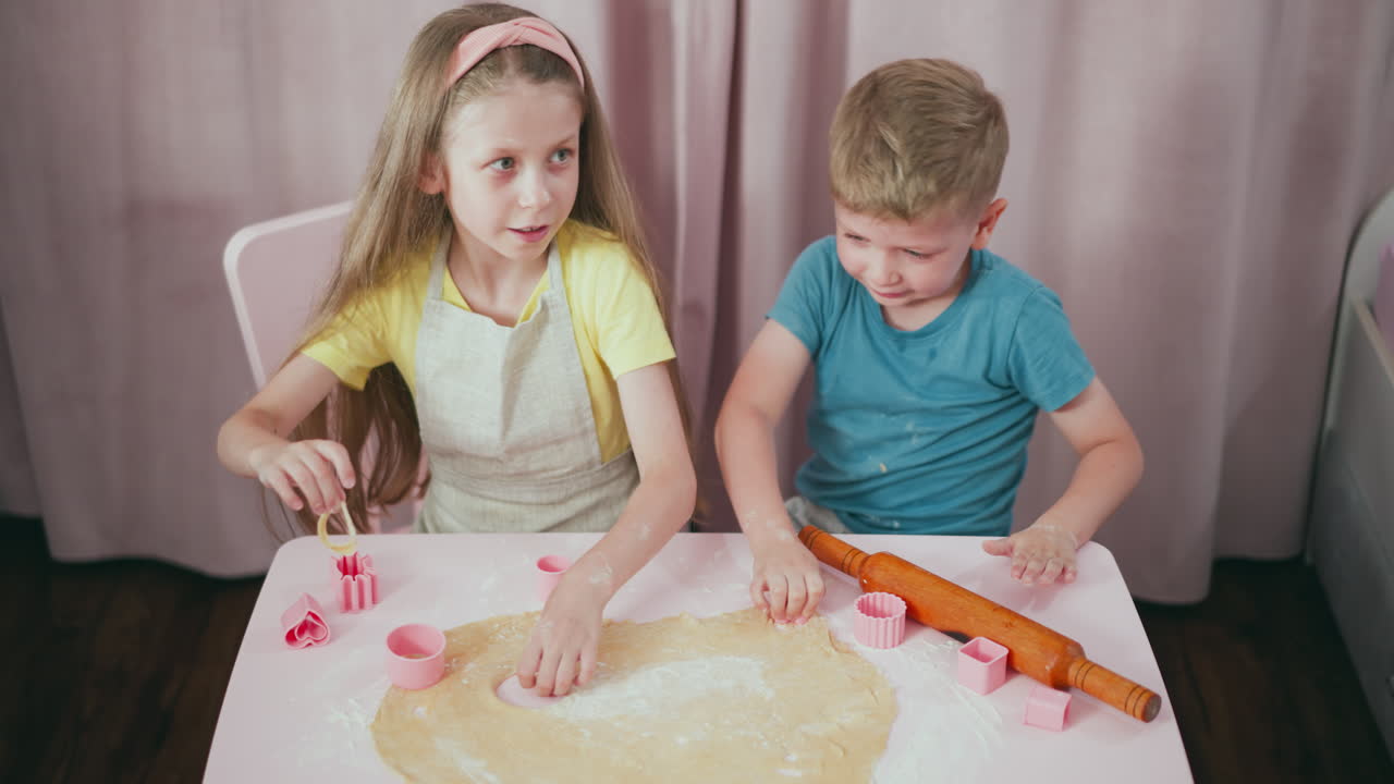 Close up of siblings making shapes with pink cutters on dough during baking session, with girl gently playing with cut out dough while boy focuses on pressing cutter into floured surface