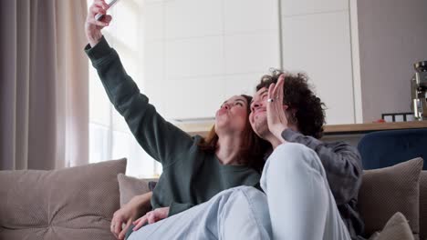 Ein-Glückliches-Brünettes-Mädchen-Macht-Ein-Selfie-Mit-Ihrem-Freund-Mit-Lockigem-Haar,-Während-Es-Zu-Hause-In-Einem-Modernen-Apartment-Auf-Dem-Sofa-Sitzt