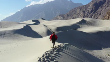 drone tracking a young happy caucasian male tourist traveling in a red jacket running the top of sand dunes in the cold desert of skardu pakistan with a mountain range in the background during day