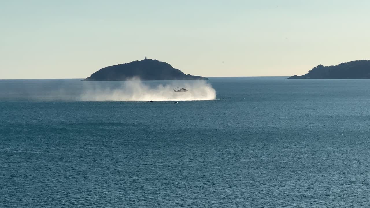 Helicopter hovering above the sea, creating dramatic spray near an island on the Ligurian coast in Italy. A seagull flies in the foreground, adding depth and motion to the scenic maritime landscape
