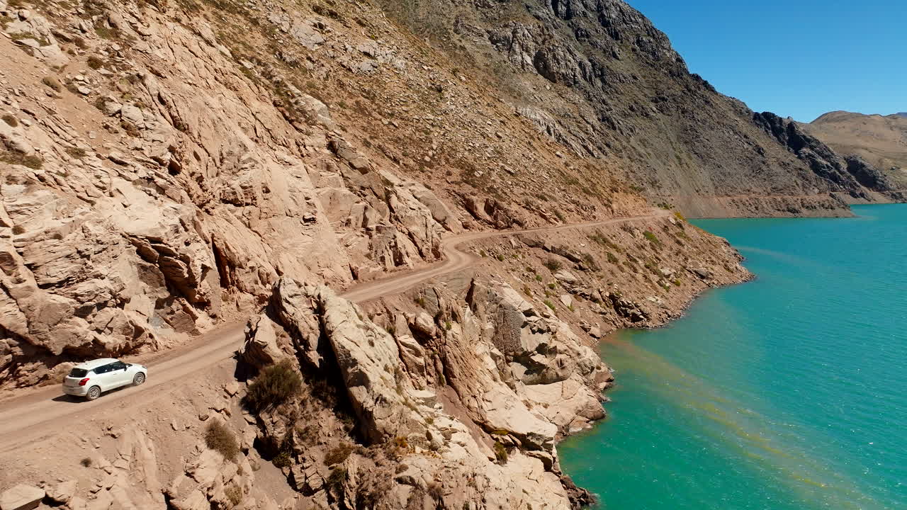 Car drive on dirt road next to blue El Yeso reservoir, Cajon del Maipo, aerial