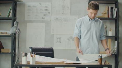 bearded man hipster standing faces to panoramic window and looks at blueprints. young architect in stylish loft office works with documents. creative architect thinking on the big drawings.