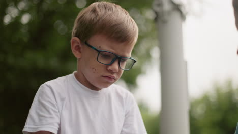 un niño con gafas parece abatido y pensativo, frunciendo el ceño ligeramente mientras mira hacia abajo, el fondo está borroso con vegetación