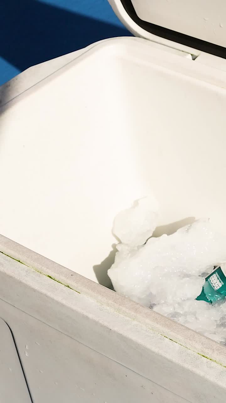 A person closes an ice box filled with ice and drinks under bright sunlight, creating a casual outdoor atmosphere