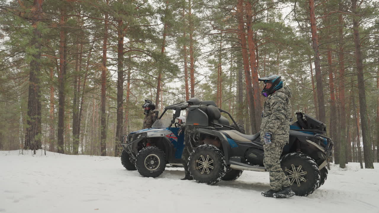 Pilota di ATV nella foresta innevata sta accanto al quad tra i pini, tuta mimetica e casco, leggera nevicata, pneumatici sulla neve compatta, in attesa di iniziare il viaggio, atmosfera calma e di attesa, preparazione all'avventura.