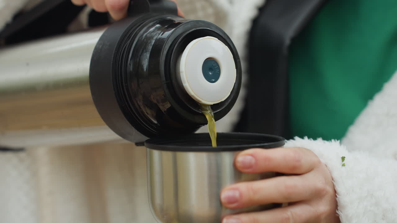 Mani che versano tè caldo da un thermos in una tazza di metallo, primo piano su presa salda e giacca di pile, vapore che sale sopra il liquido caldo, maglietta verde visibile, sfondo di parco autunnale, escursionista che si prende una pausa per riscaldarsi