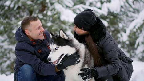 a man and a woman sitting hugging a dog siberian husky in the winter forest smiling and looking at each other and at the camera. slow motion happy family