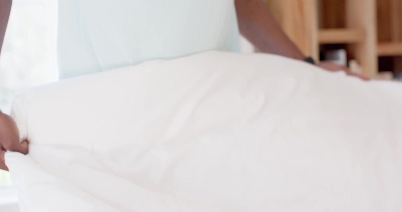 Man is grasping duvet corner, lifting and spreading duvet on bed near window, tidying bedding