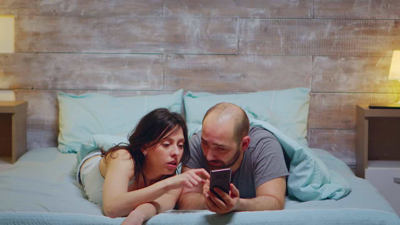 Husband and wife using phone while laying in bed