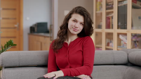 Confident-young-brunette-woman-sitting-on-living-room-couch-looking-at-camera-smiling