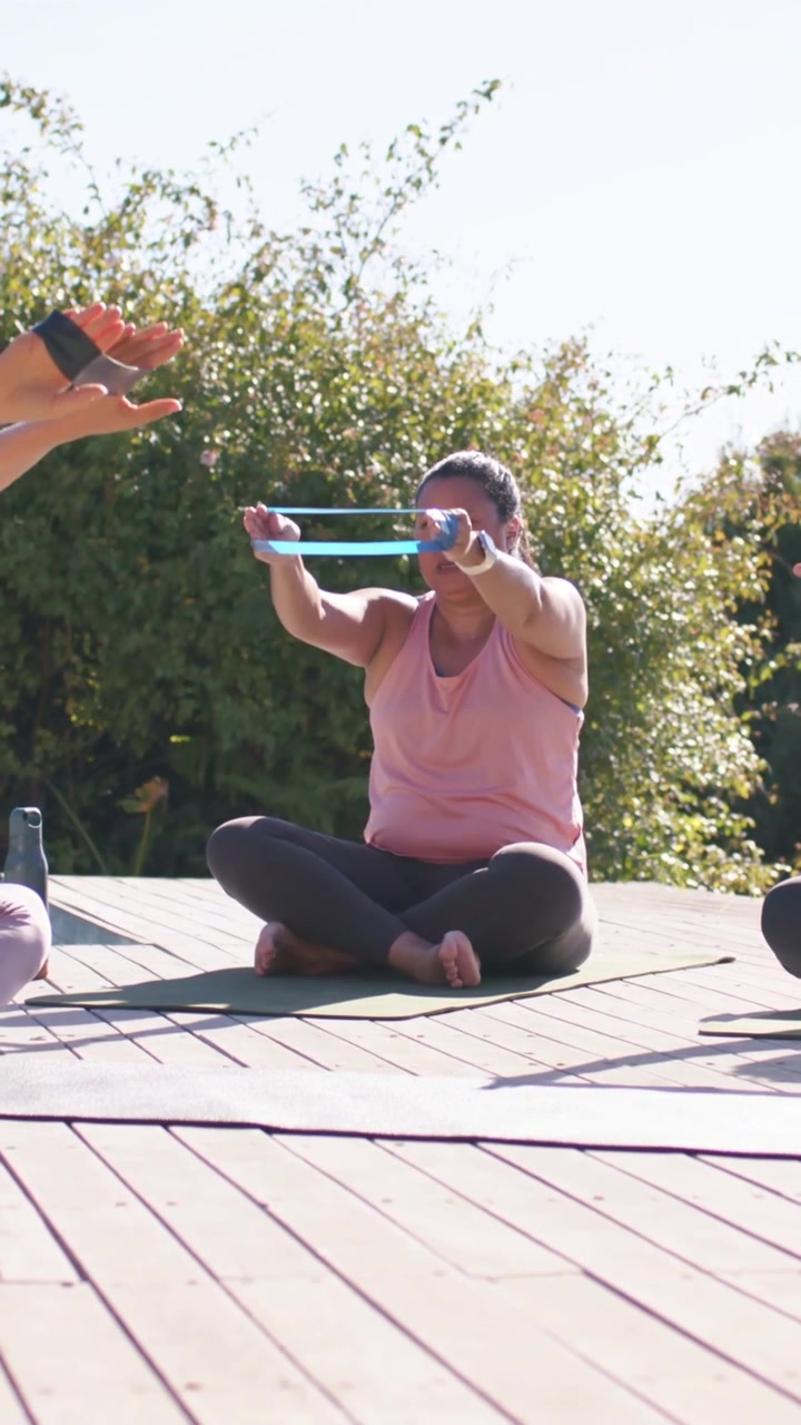 Video vertical: Mujer siguiendo al instructor, levantando una banda azul en una colchoneta verde en la terraza, con top rosa.