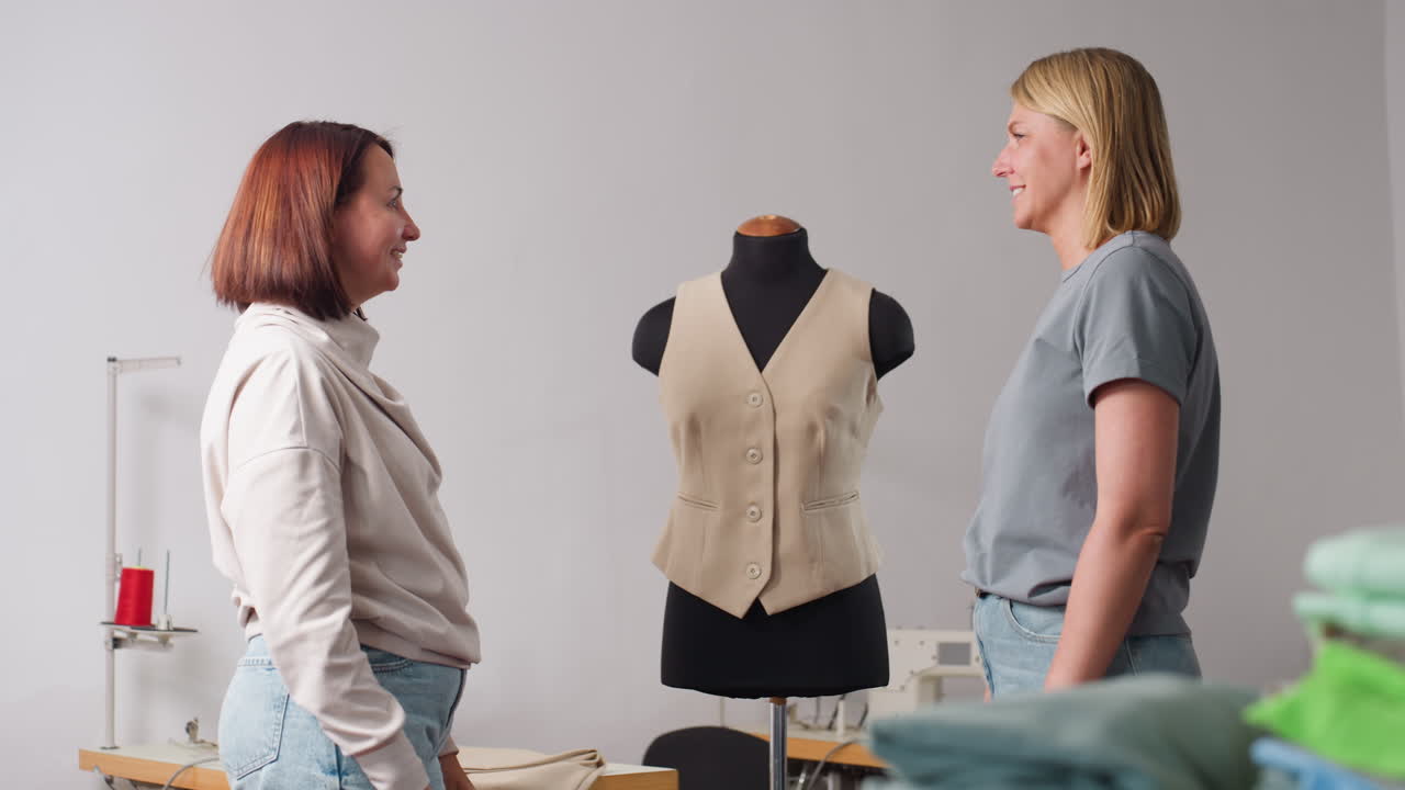 Stylists exchange high five beside beige vest on black mannequin, grinning proudly in sewing studio with machines and folded fabrics, celebrating successful design collaboration inside workspace