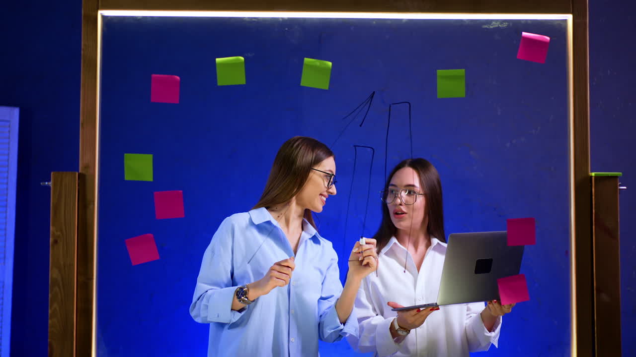 Brunette in glasses and white shirt drawing graphs on the glass board. Girl standing beside is holding a laptop. Business strategy and planning.