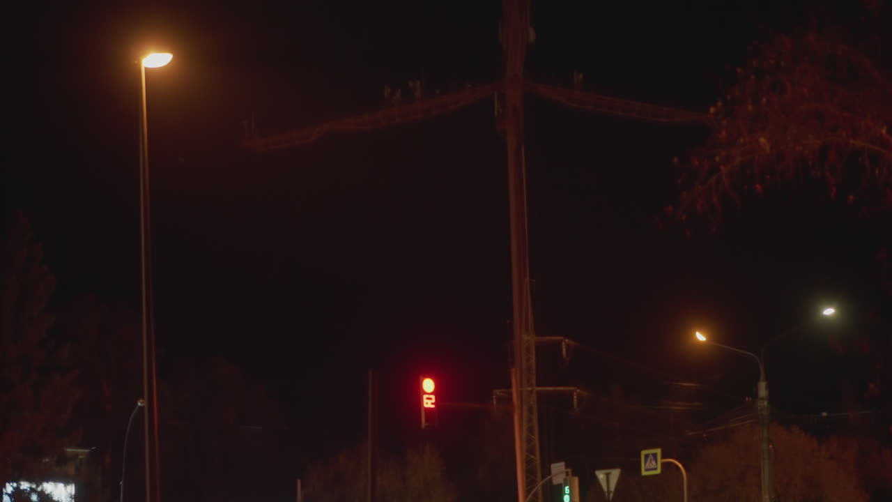 night intersection with red signal. Empty urban road under dim lamp glow, tall utility pole silhouette against dark sky. Red traffic light dominating scene, faint neon from distant signage, reflective