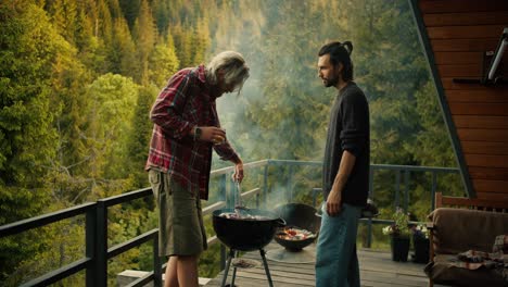 dos chicos freír salchichas en la parrilla en la grilla. un hombre con una camisa roja da la vuelta salchicha usando pinzas especiales contra el telón de fondo de las montañas y el bosque