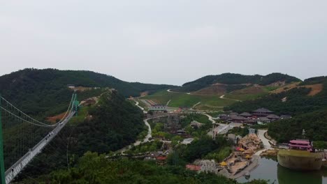 Aerial-ascending-view-behind-trees-revealing-suspension-glass-bridge-and-beautiful-scenery-at-Huaxiacheng-theme-park-in-Weihai,-China