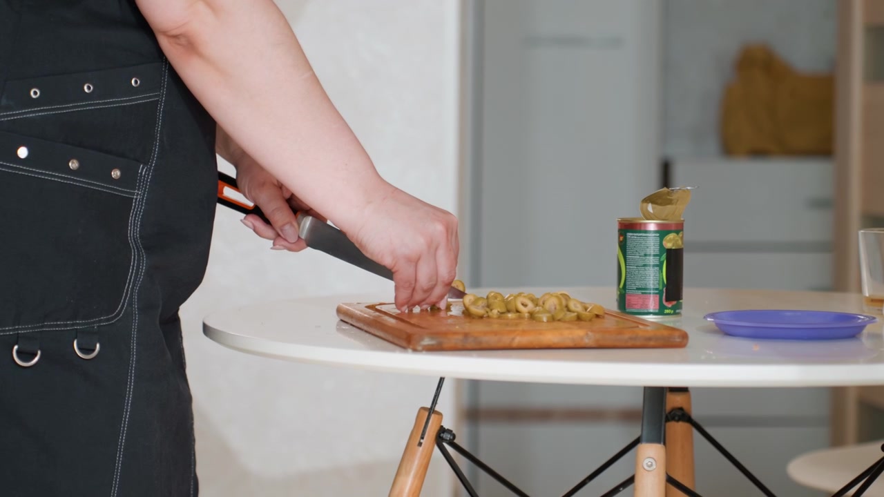 Tampilan dekat tangan menggunakan pisau untuk mengiris sayuran segar di talenan kayu di samping meja bundar putih, menunjukkan persiapan makanan yang aktif, penataan dapur yang rapi, cahaya alami, dan masakan rumahan yang santai