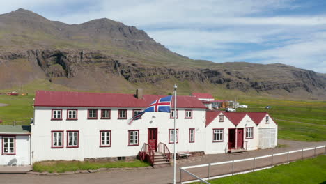 Orbit-drone-view-around-flag-fluttering-in-the-wind-out-of-public-office-in-Icelandic-countryside.-Aerial-view-Iceland-flag-waving-out-of-a-typical-house-in-icelandic-highlands