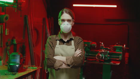 portrait of young female welder in apron and goggles putting on the protective mask and looking at camera at work in workshop