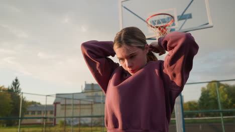 woman tying hair backward outdoors with basketball hoop and urban building in background, natural sunlight highlights her focused expression as she prepares for sports