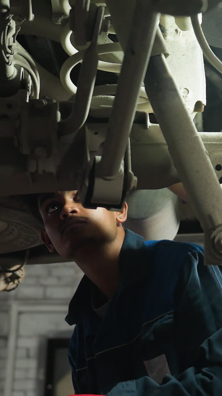ingeniero indio trabajando debajo de un coche, apretando un perno con expresión enfocada, la luz se refleja en su cara, usando guantes y uniforme azul, ocupado en la reparación de automóviles dentro de un garaje profesional