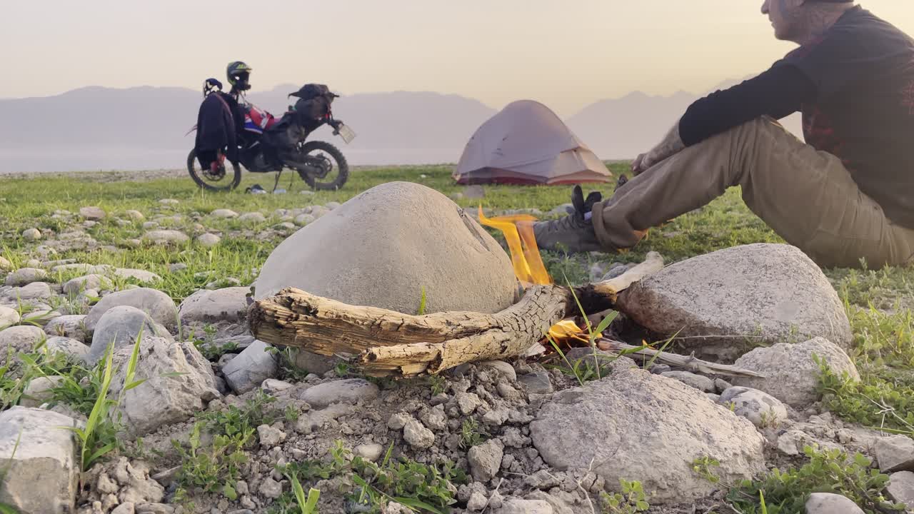 Mototurismo d'avventura: un viaggiatore siede accanto a un falò vicino a una tenda e una moto dual-sport al tramonto, con uno splendido paesaggio montano e un lago sullo sfondo.