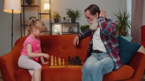 Smiling-senior-grandfather-playing-chess-board-game-with-teen-granddaughter-child-girl-kid-at-home