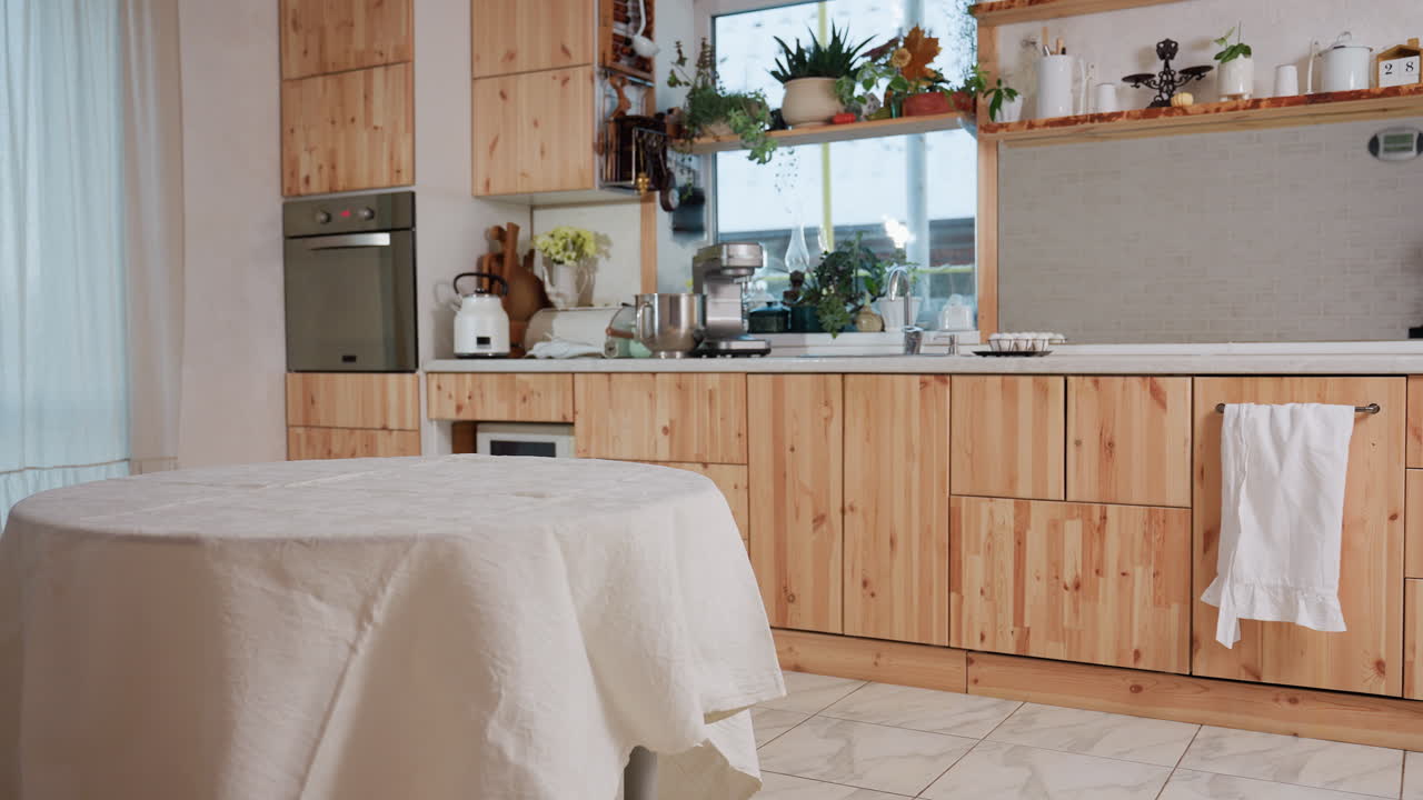 View of modern kitchen interior with wooden cabinets, countertop appliances, indoor plants, and cozy dining area with table covered in white cloth near large window