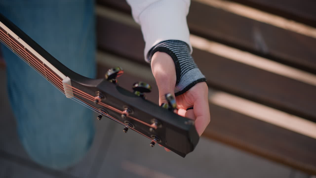 primo piano di un chitarrista caucasico che accorda una chitarra acustica su una panchina di legno, la mano sinistra con il guanto che regola i piroli della paletta, luce solare calda e ombre del parco, preparazione meticolosa per l'esibizione, dettaglio intimo