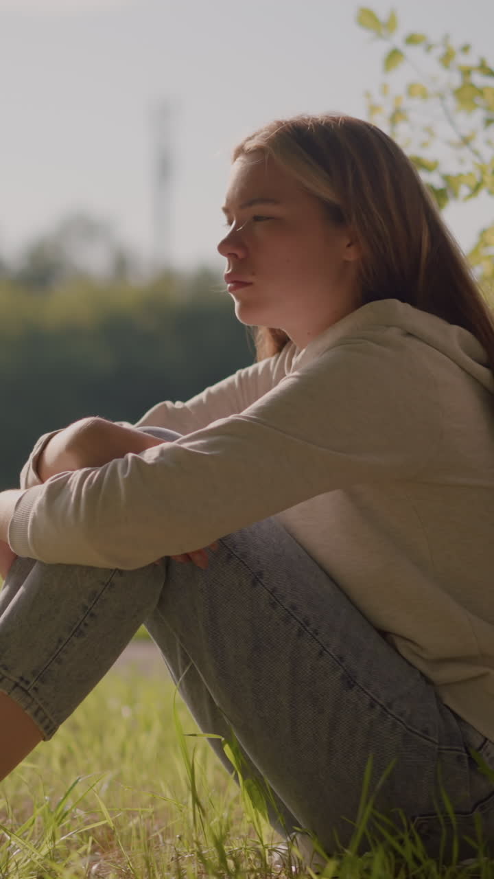Close-up side view of young woman seated thoughtfully on grassy ground, she leans forward with arms folded over knees, soft blur in background includes stadium poles and greenery