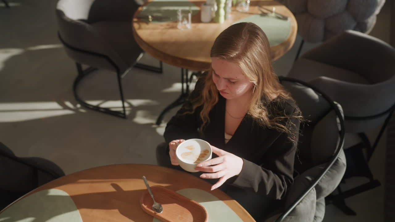 Giovane donna in un caffè illuminato dal sole che ammira la schiuma del latte, una fotografa freelance si ferma tra uno scatto e l'altro per studiare la latte art e l'aroma caldo, luce solare che proietta ombre morbide sul tavolo di legno, tazza di ceramica, piattino