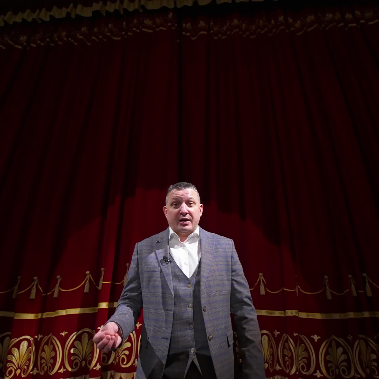 Theater host on red curtains background. Man in grey costume talking on a big stage. Portrait of an actor performing on stage