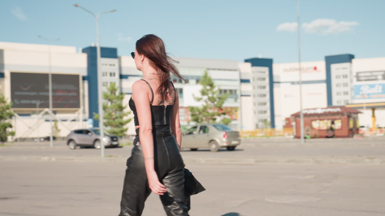 Lady in black singlet and leather pants walking confidently on urban street holding jacket in right hand, sunglasses on, attitude strong, background with cars, buildings, trees, and clear blue sky