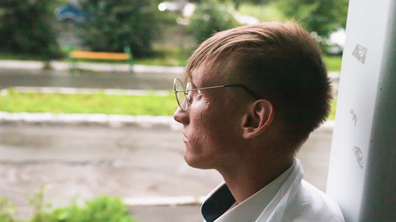 Close view of young botany student in white coat and glasses leaning against wall during outdoor break, reflecting calmly in natural urban environment with greenery after sipping juice