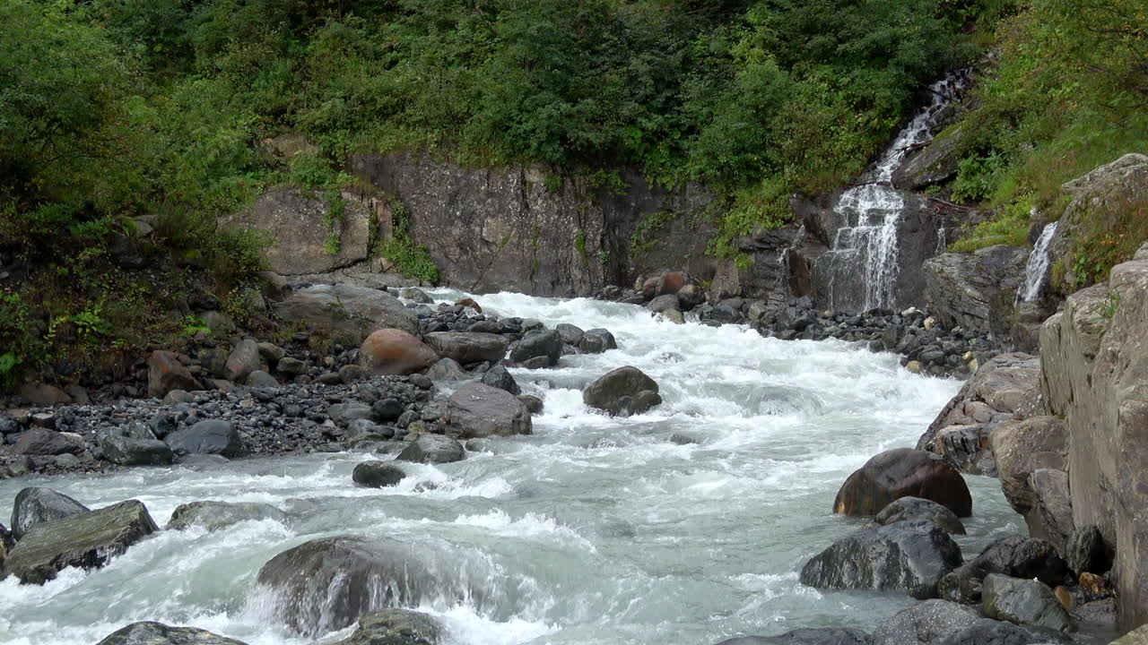 Sungai pegunungan yang indah dengan jeram yang deras mengalir melalui ngarai berbatu dan hutan hijau, dengan air terjun kecil di sebelah kanan, difilmkan di Dombay, Kaukasus, Rusia