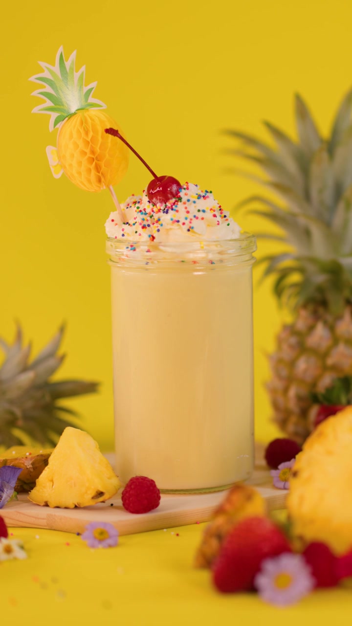 Una mano aggiunge una decorazione all'ananas a un frullato cremoso con panna montata, ciliegia e codette, illuminazione brillante