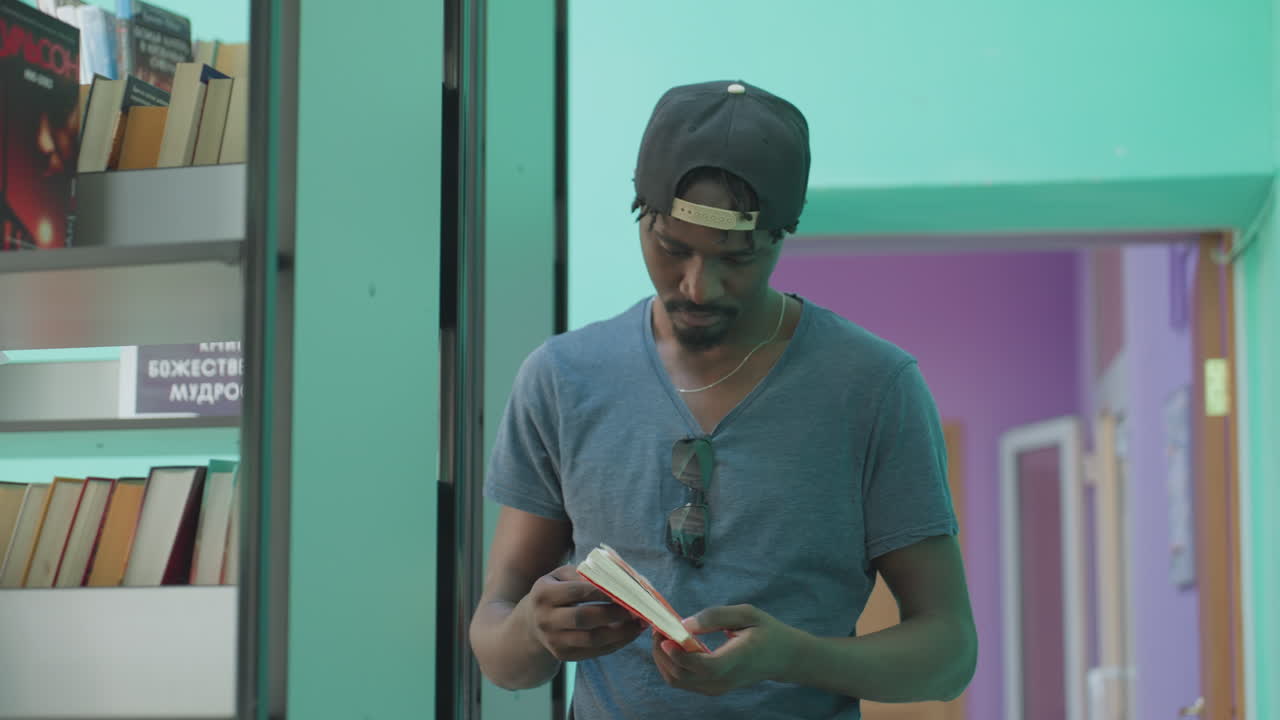 Focused man in casual shirt and cap reaches for book on library shelf, flipping pages as sunglasses reflect surroundings, with vibrant teal wall and purple hallway in soft background