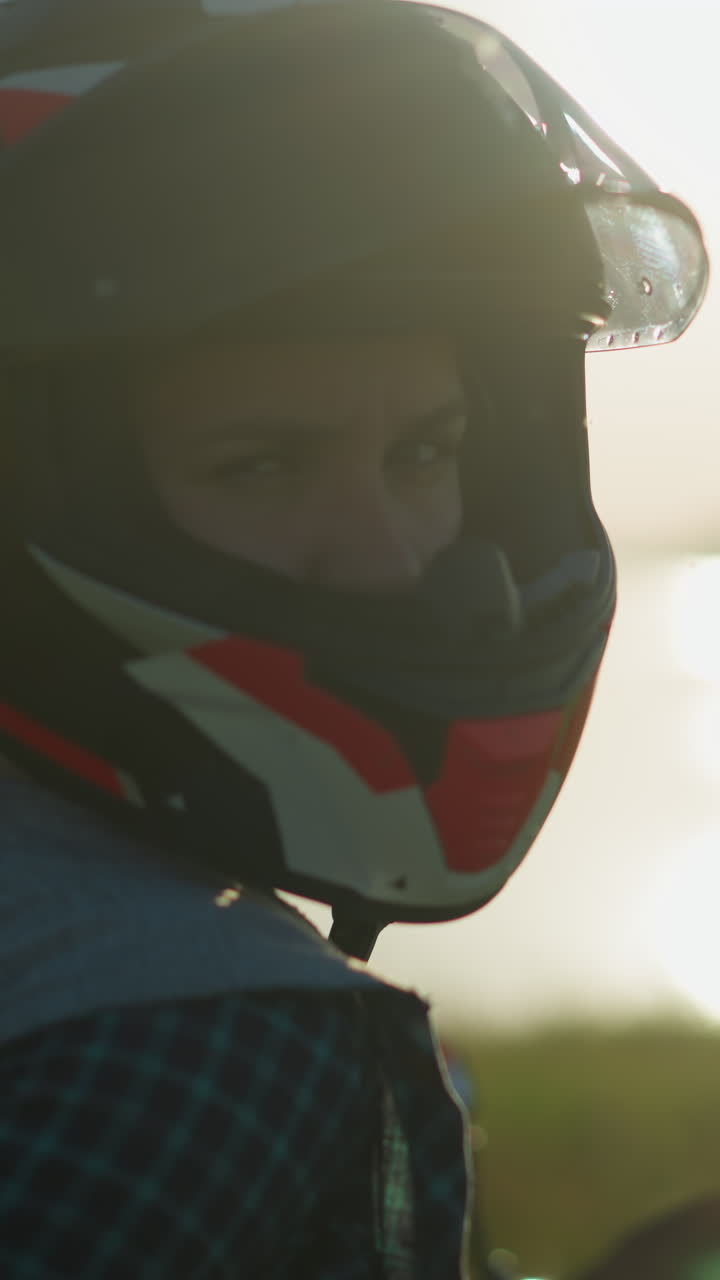 una mujer con un casco se sienta en una motocicleta en un campo de hierba, mirando hacia la cámara, con una vista borrosa del cuerpo de agua en la distancia
