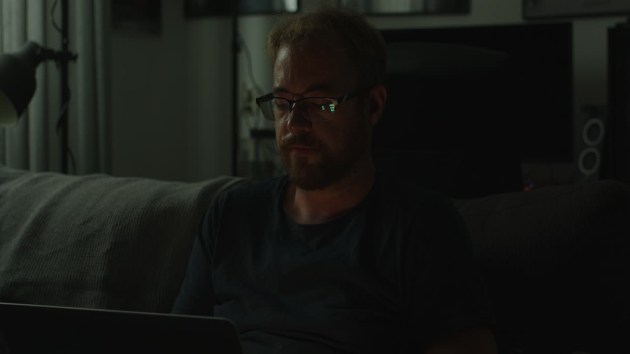 A man works on his laptop in a very dark living room, he turns his head towards a noise