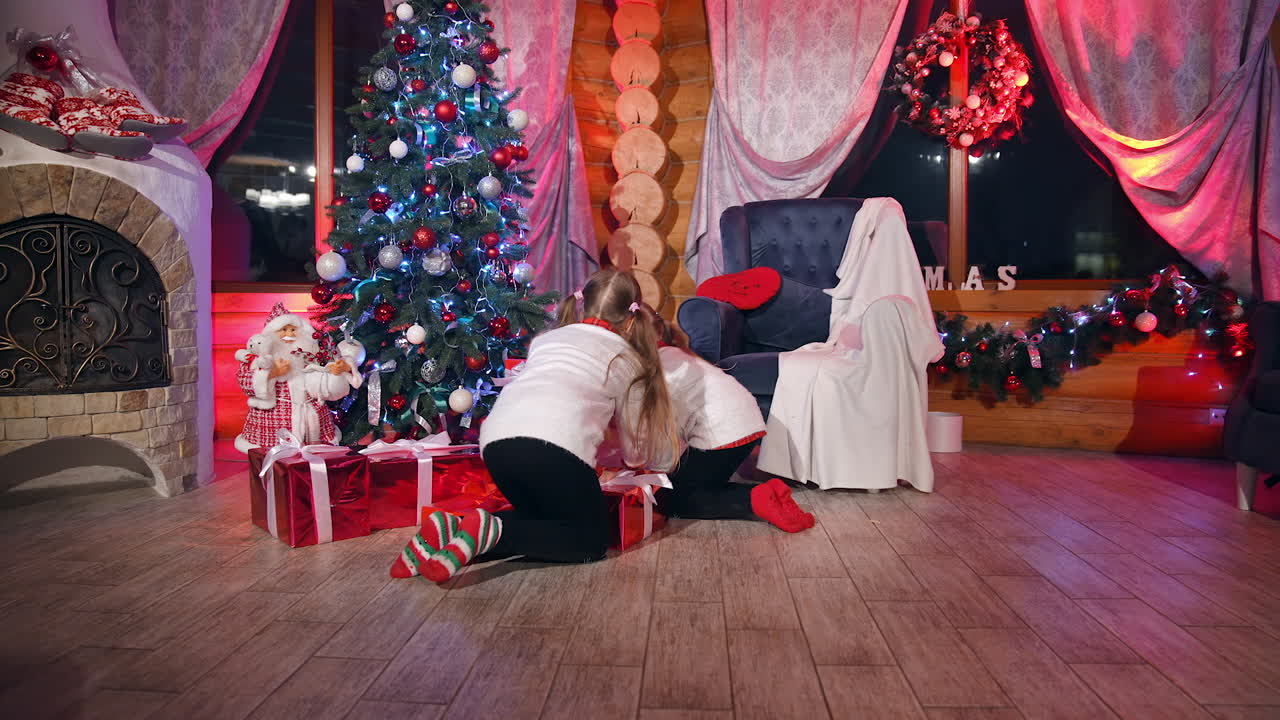Children in Christmas decorated room. Little girls coming to a Christmas tree with many gift boxes and taking cards. New Year eve.