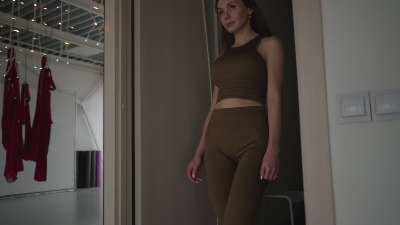 Well lit gym studio with minimal modern interior and equipment as female athlete dressed in two piece brown cloth walks through doorway confidently, conveying strength focus and wellness