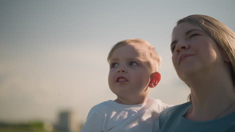 close-up of a woman in a blue dress carrying her young son, who is wearing a white top. the boy is attentively pointing at the surroundings, a moment of curiosity and exploration on a sunny day