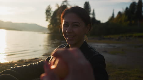 lady gives apple to boyfriend on riverbank. young woman offers fruit to partner silhouette resting near lake. couple enjoys romantic atmosphere at sunset