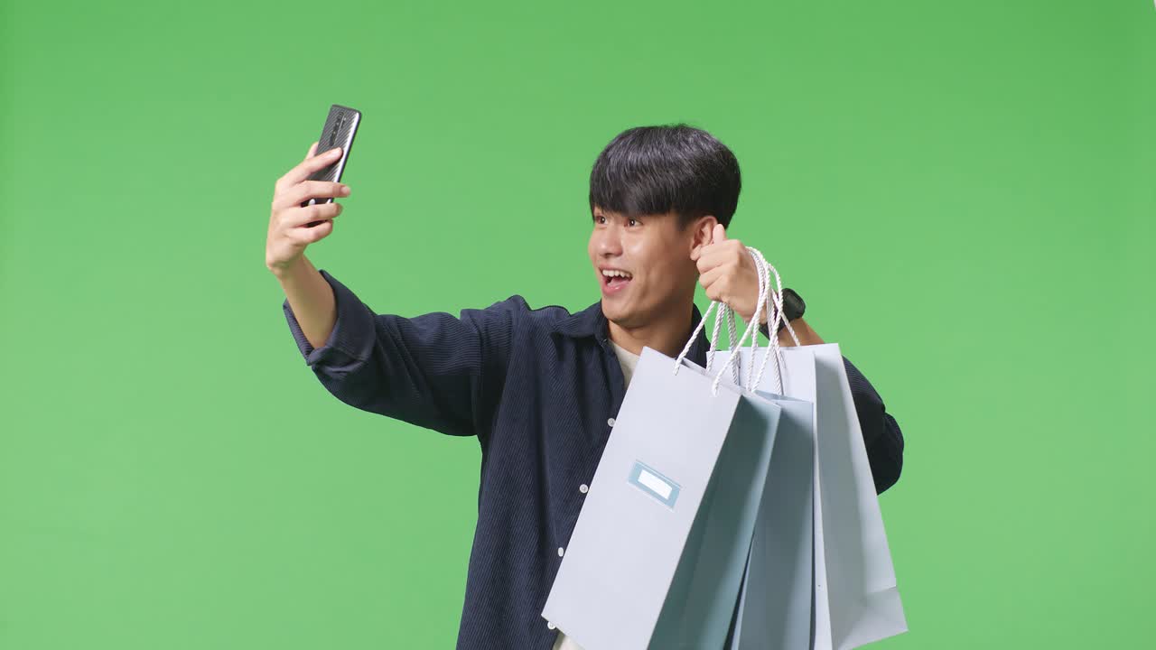 초록색 화면 앞에 서 있는 동안 휴대 전화로 비디오 통화를 하는 쇼핑 가방 을 가진 아시아 쇼핑 남자